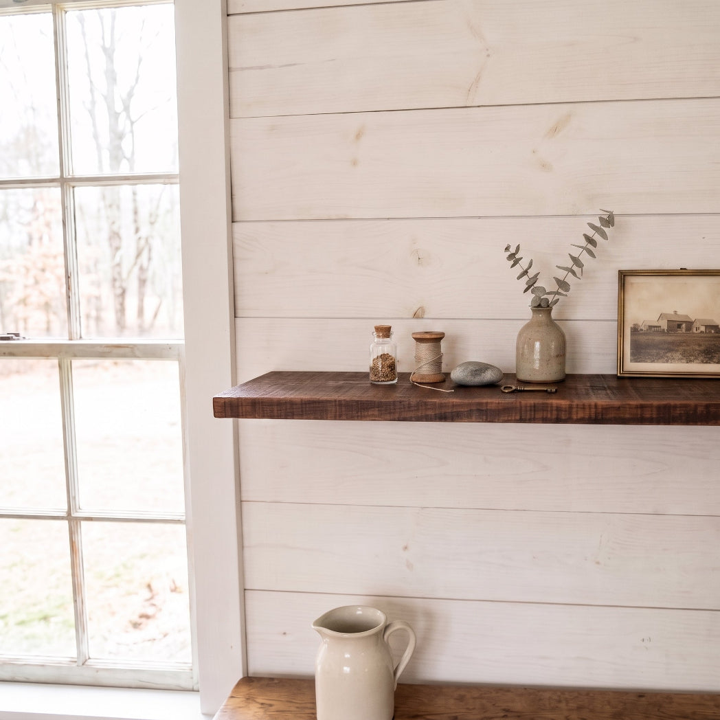 A rustic dark wood floating shelf mounted on a white shiplap wall, styled with small glass jars, a ceramic vase with eucalyptus, a stone, and a framed print. A white ceramic pitcher sits below on a lower shelf, next to a window looking out onto a wooded yard.