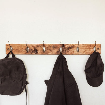 Wooden coat rack with hooks holding a backpack, jacket, and cap against a white wall.