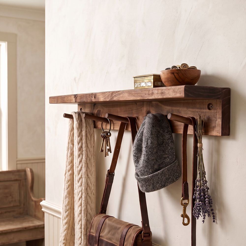 A rustic wooden wall-mounted shelf with hanging hooks, styled in a home entryway with a cream cable-knit scarf, keys, a gray knit beanie, a brown leather shoulder bag, and dried lavender.