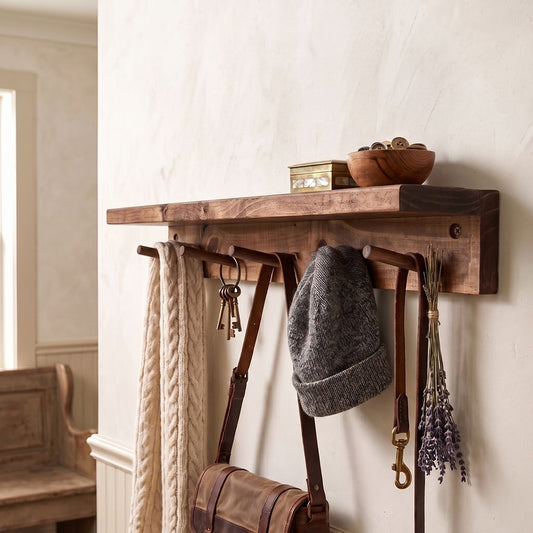 A rustic wooden wall-mounted shelf with hanging hooks, styled in a home entryway with a cream cable-knit scarf, keys, a gray knit beanie, a brown leather shoulder bag, and dried lavender.
