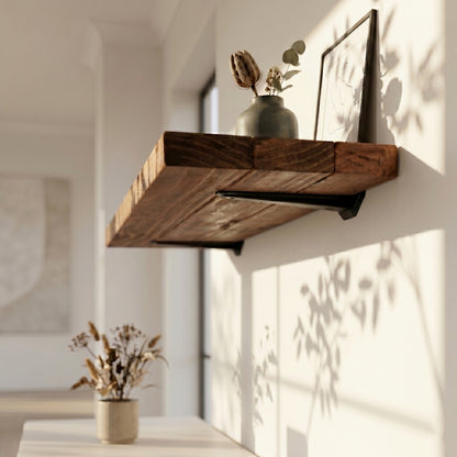 Low-angle side view of a chunky rustic pine floating shelf in walnut brown, mounted on a white wall with black metal brackets. The timber ledge is styled with a small ceramic vase holding a dried flower and a leaning picture frame, featuring soft leaf shadows cast by natural sunlight.