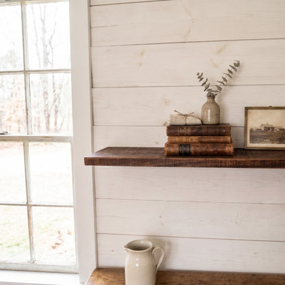 Front straight-on view of a rustic solid wood floating shelf styled with antique books and a small vase, mounted on white shiplap walls next to a bright window.